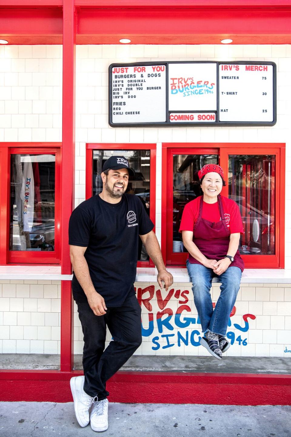 A bearded man in a T-shirt and ballcap stands next to an aproned woman sitting on the counter outside a hamburger stand.