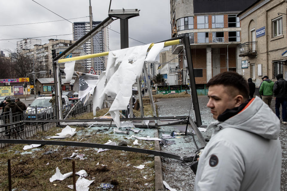 A young man in a weatherproof jacket stands in front of a framelike structure from which a shredded screen is hanging.