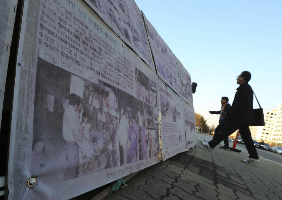 In this April 2, 2019, photo, people walk past a photo of guards unloading children from a truck at the Brothers Home in Busan, as it is displayed in front of National Assembly in Seoul, South Korea. A notorious South Korean facility that kidnapped, abused and enslaved children, adults and the disabled for a generation was also shipping children overseas for adoption, part of a massive profit-seeking enterprise that thrived by exploiting those trapped within its walls, The Associated Press has found.(AP Photo/Ahn Young-joon)