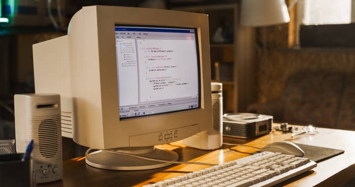 Retro computer with code on the screen sitting on a wooden desk, surrounded by speakers, a keyboard, and various electronic components