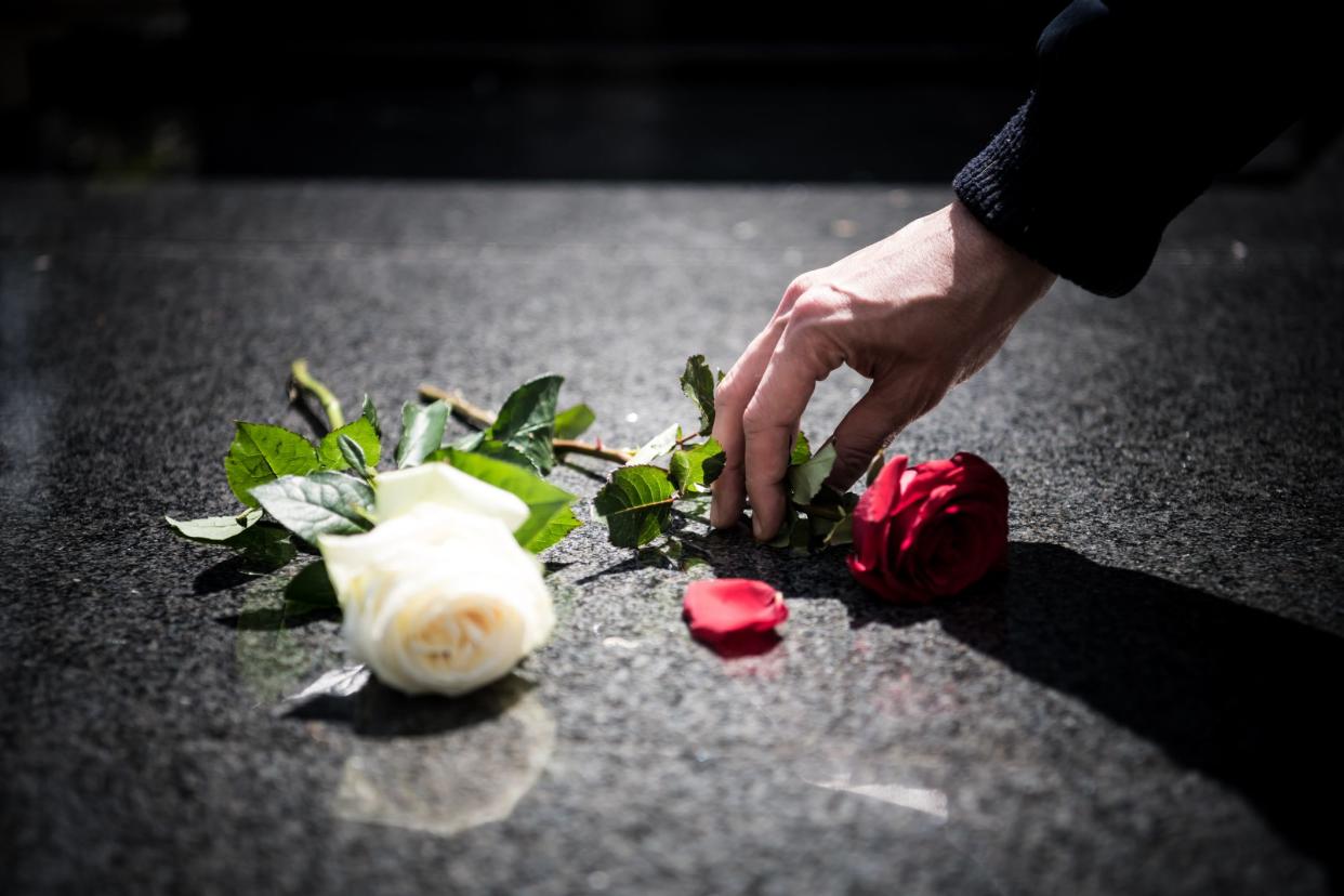 A man's hand places a red rose with stem on a horizontal granite gravestone next to a white rose with stem on the left, surrounded by dark shadowing