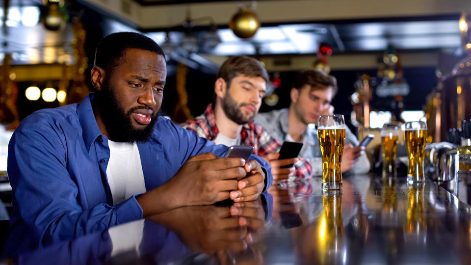 Sad multiethnic men surfing internet on phones in pub instead of communicating.