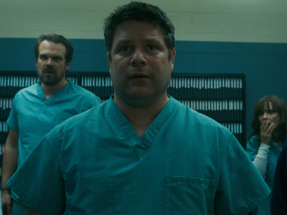 sean astin as bob in stranger things, wearing scrubs and standing in front of dr. owens, hopper, joyce, and mike in a room in hawkins lab in stranger things