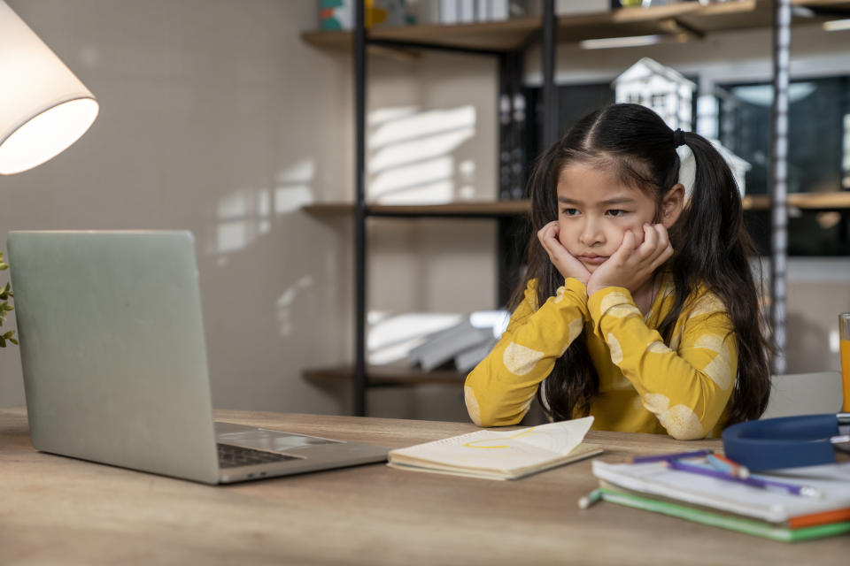 Young girl with her chin in her hands looking bored in front of a laptop, with books on the table