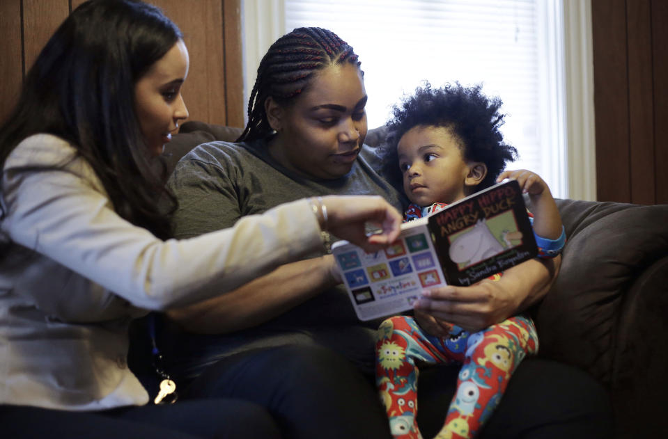 In this Feb. 3, 2014 photo, caseworker and home visitor Stephanie Taveras, left, reads a book with Ashley Cox, center, and Cox's 16-month-old son Jaiden, right, at the family's home in Providence, R.I. The city has begun an effort to boost language skills for children from low-income families by equipping them with audio recorders that count every word they hear. During home visits, social workers go over the word counts with parents and suggest tips to boost the child’s language skills. (AP Photo/Steven Senne)
