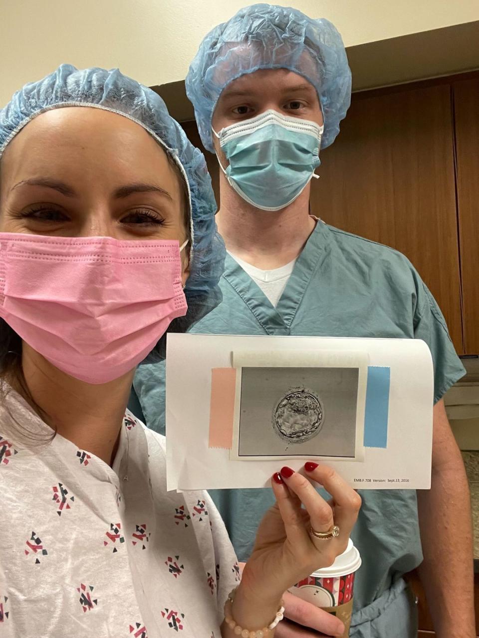 a photo of aubrey in a mask holding up a photo of the embryo with her husband in a mask behind her