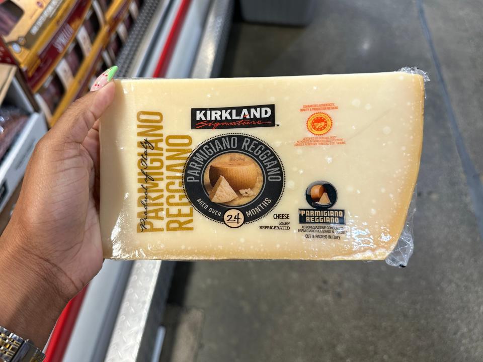 A hand holds a large piece of Kirkland Signature Parmesan cheese with a black label and an image of a cheese wheel on the packaging