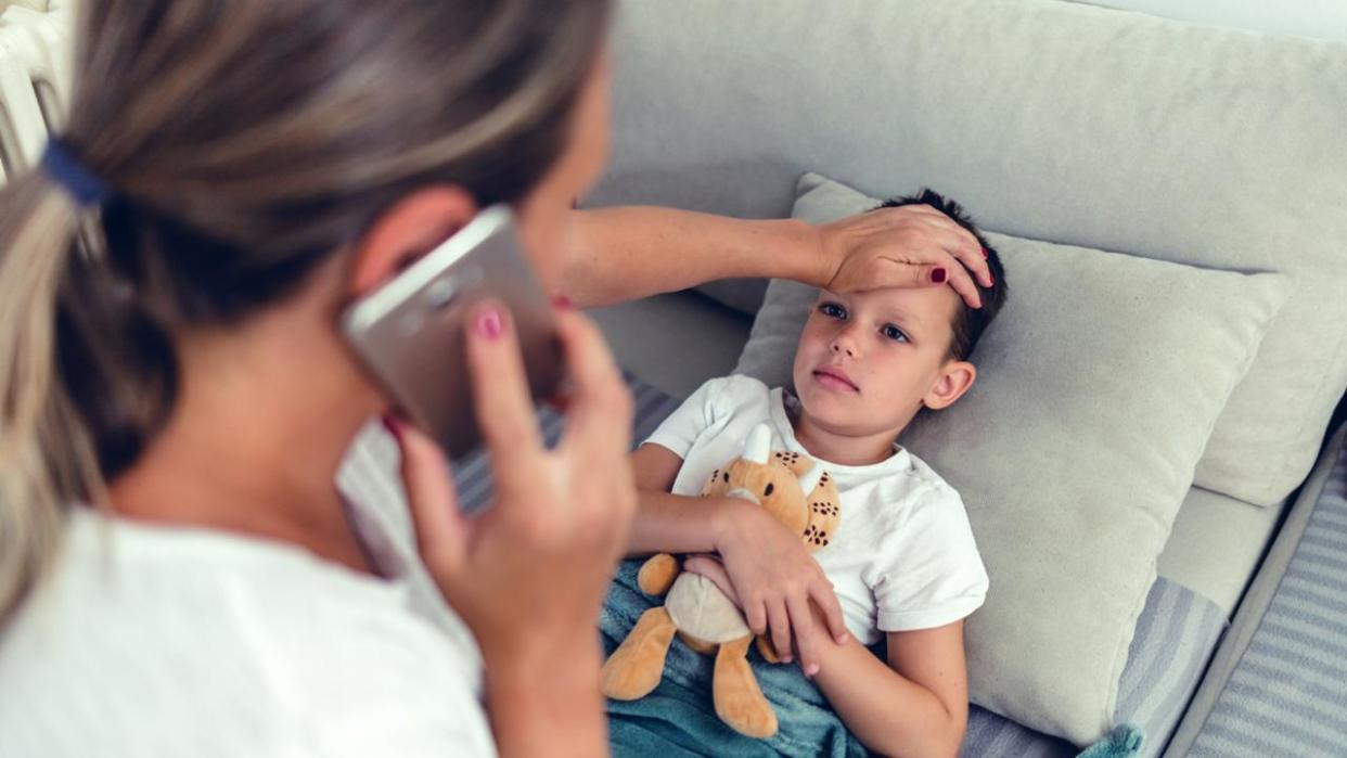 Photo of worried mother checking temperature of her little son who is lying in bed with fever. Young blond hair mother checking the temperature of her ill 6 years old boy on a couch in the living room at home, calling doctor.