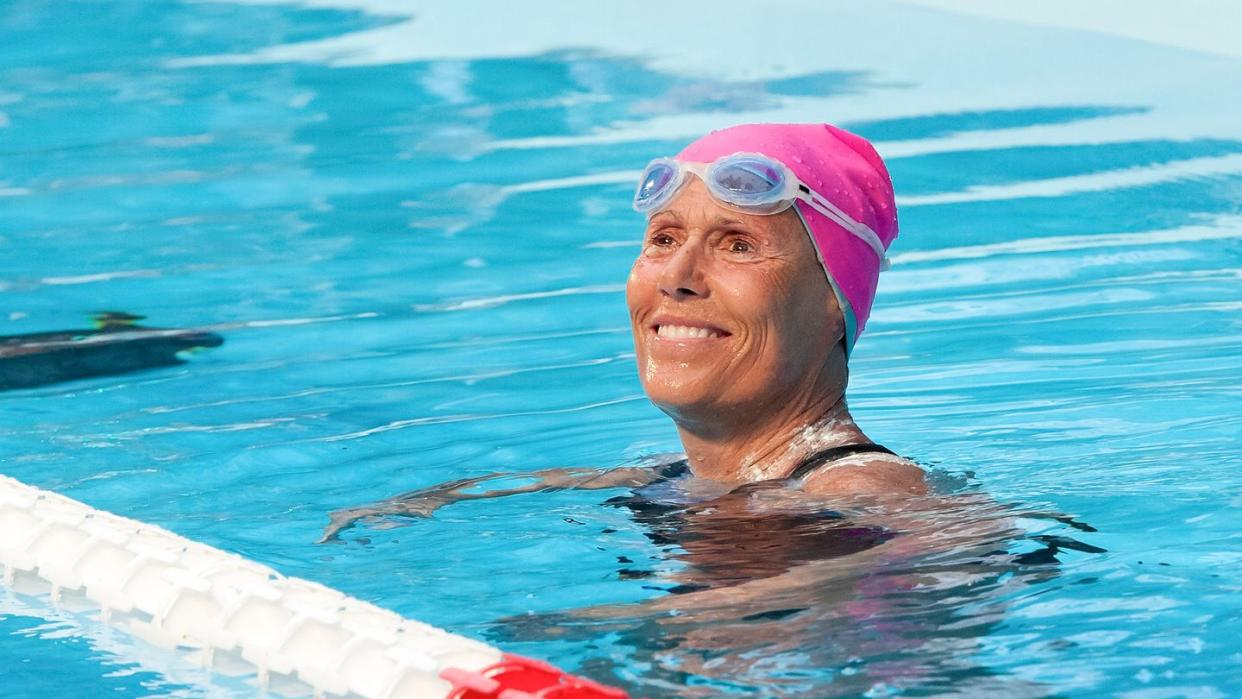 diana nyad floats in a swimming pool and looks past the camera smiling, she wears a pink swimming cap and white and blue googles on her forehead