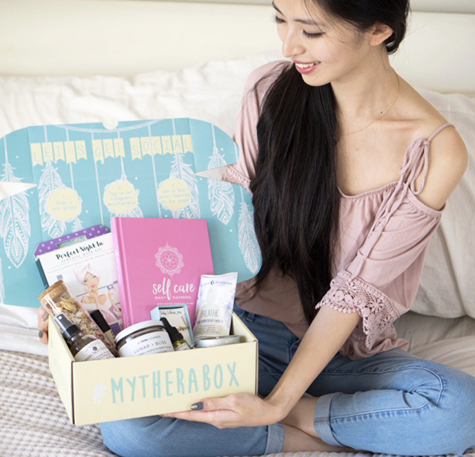 woman sitting on bed in blue jeans and pink shirt holding open gift box TheraBox (photo via My TheraBox)