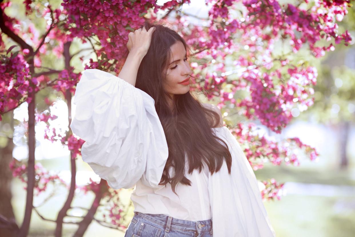 Happy woman in spring. Elegant dreaming woman wearing white cotton shirt with wide sleeves lanterns flounces, posing near pink blossom trees, touches long dark hair. Fashion model Outdoor portrait