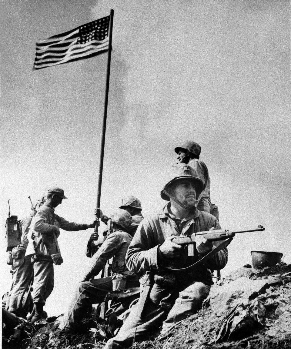 A U.S. Marine from the 5th Division of the 28th Regiment stands guard atop Mt. Suribachi at Iwo Jima in the Volcano Islands of Japan as others hoist the American flag during World War II, Feb. 23, 1945. (Louis R. Lowery / U.S. Marine Corps via AP file)