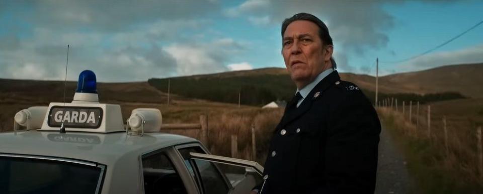 ciaran hinds in the land of saints and sinners movie, his character wears a police uniform and looks thoughtful as he stands in front of some fields