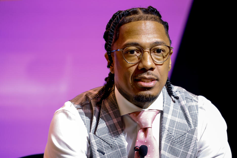 Nick Cannon in a white shirt, checkered vest, and pink tie, with braided hair, speaks at an event