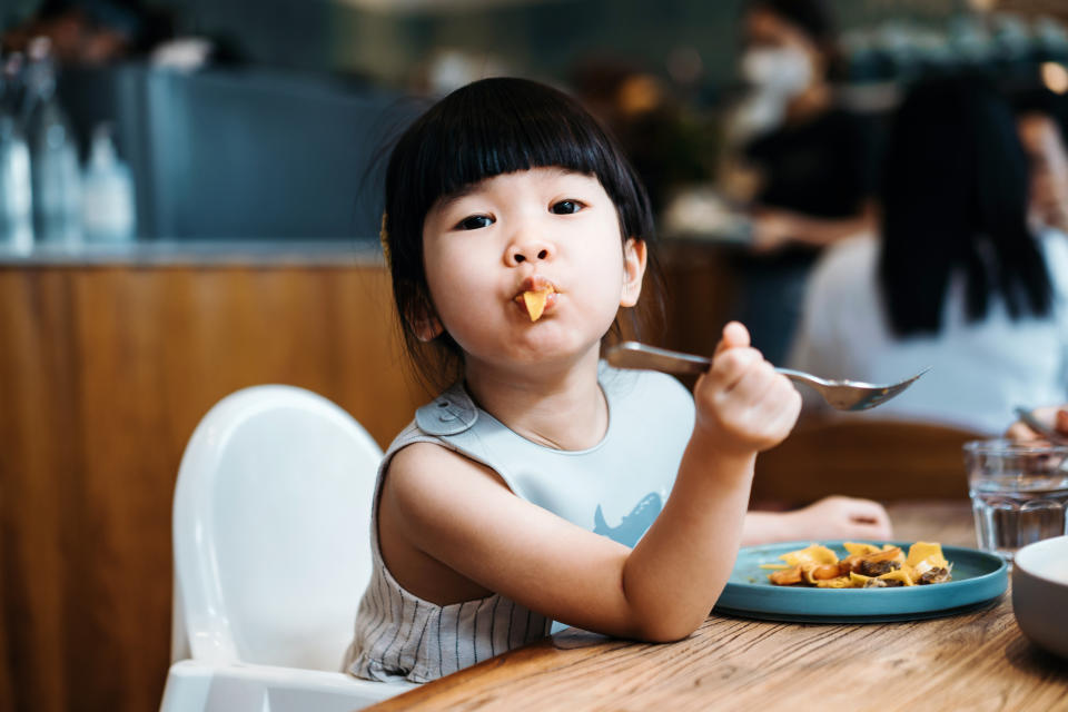 Portrait of a little girl sitting on a high chair having pasta for lunch in cafe. She is making a face while looking at camera sitting at dining table. People, food and lifestyle concept