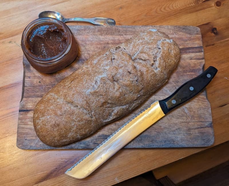 loaf of bread on wood cutting board with knife and jar of jam