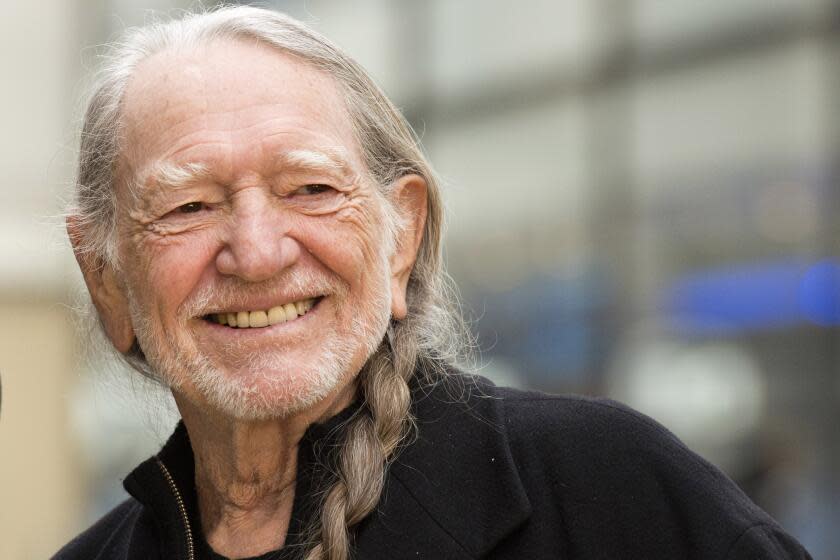 Willie Nelson, with a braid in his grey hair, smiles against an out-of-focus background