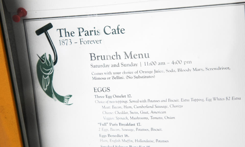 The menu for the Paris Cafe hangs in its front window, Tuesday, Aug. 4, 2020 in New York. Many iconic bars, dinners and restaurants have closed or are teetering towards closing due to the coronavirus pandemic. (AP Photo/Mark Lennihan)