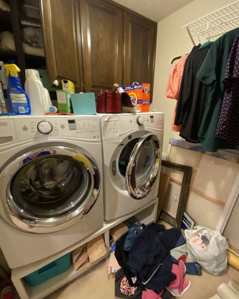 Wood cabinets with washer and dryer below, messy top with detergent and cleaning supplies everywhere, hanging clothes on rack, pile of clothes on floor