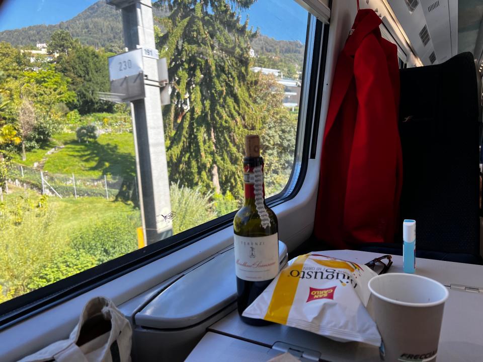 a bottle of wine and a bag of chips on a table in front of a train seat on a European train passing lush trees