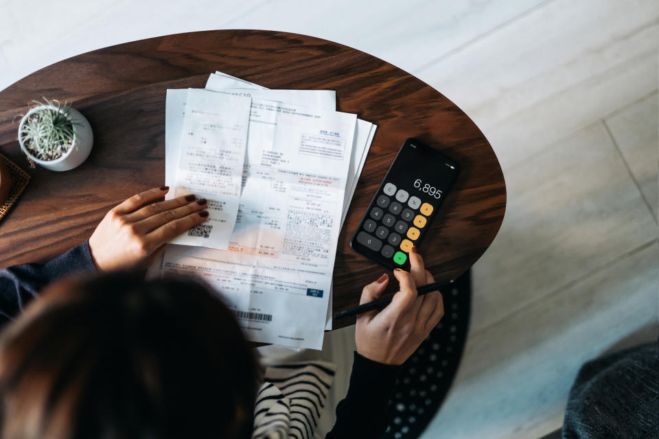 a woman's left hand rests on a stack of papers with numbers; her right hand plugs something into a calculator