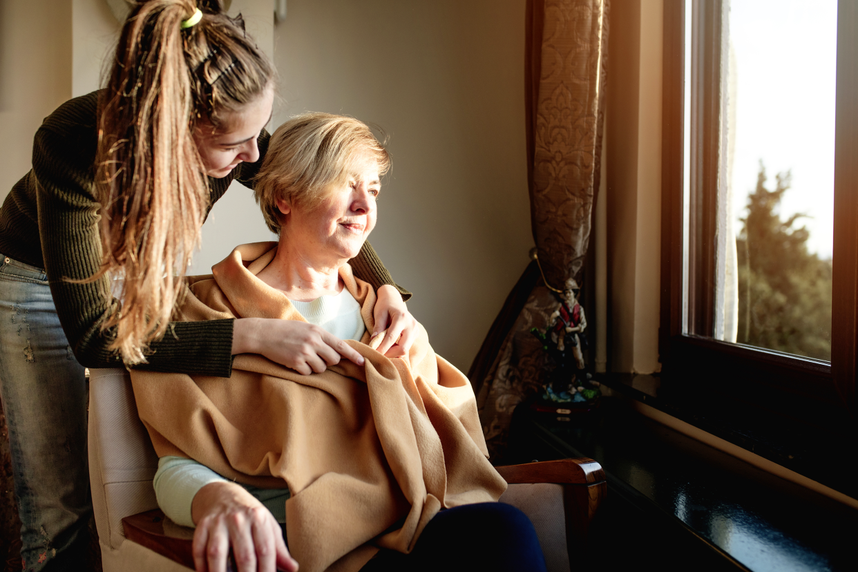 Teenage granddaughter putting beige blanket over grandmother with dementia, grandmother is sitting in a chair facing and looking out of the window, wood on window sill, beige wall in the background