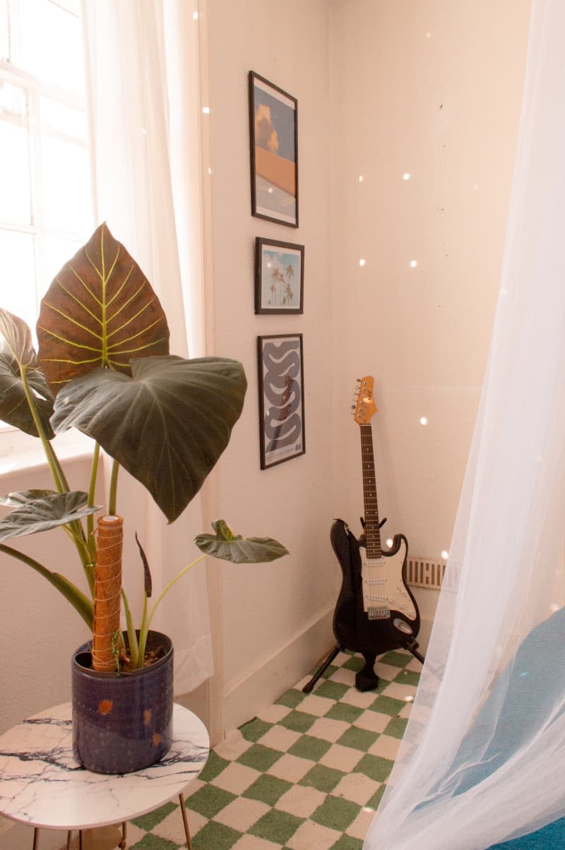 Three framed art pieces hang above a guitar in the corner of a bedroom.