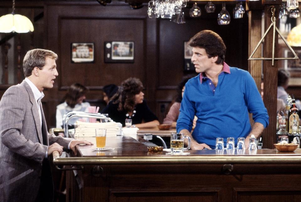 Two men, Woody Harrelson and Ted Danson, talk at a bar with drinks and glasses in front of them in a scene from the TV show "Cheers." The bar is busy