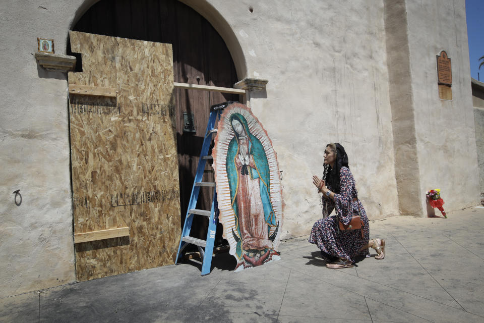 Alecia Ballin kneels in prayer in front of the boarded up entrance to the fire-damaged San Gabriel Mission Sunday, July 12, 2020, in San Gabriel, Calif. A fire on Saturday destroyed the rooftop and most of the interior of the nearly 250-year-old California church that was undergoing renovation. (AP Photo/Marcio Jose Sanchez)