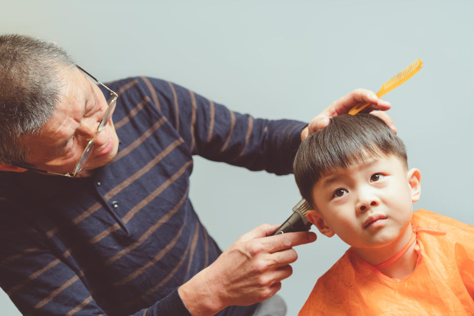 Man wearing glasses and a striped sweater uses clippers to give a young boy in an orange cape a haircut