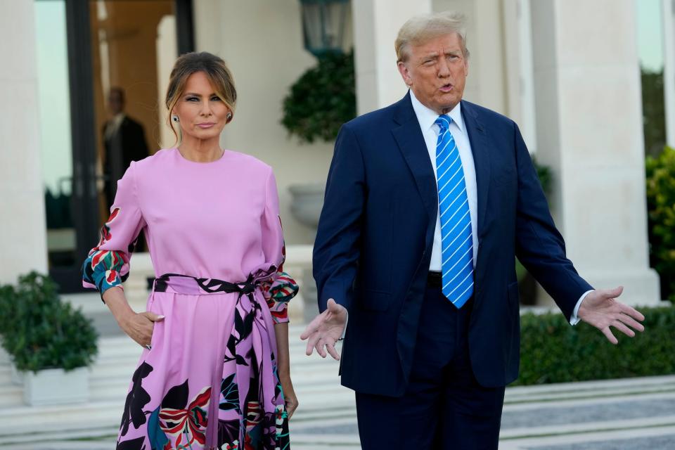Melania Trump, in a pink dress long sleeve dress, and Donald Trump, in a blue suit and striped tie, speaking with reporters in front of a white mansion