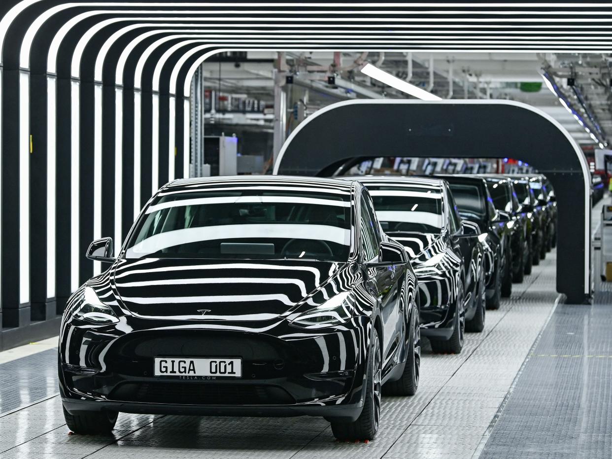 A row of Tesla cars leave a factory surrounded by lights.