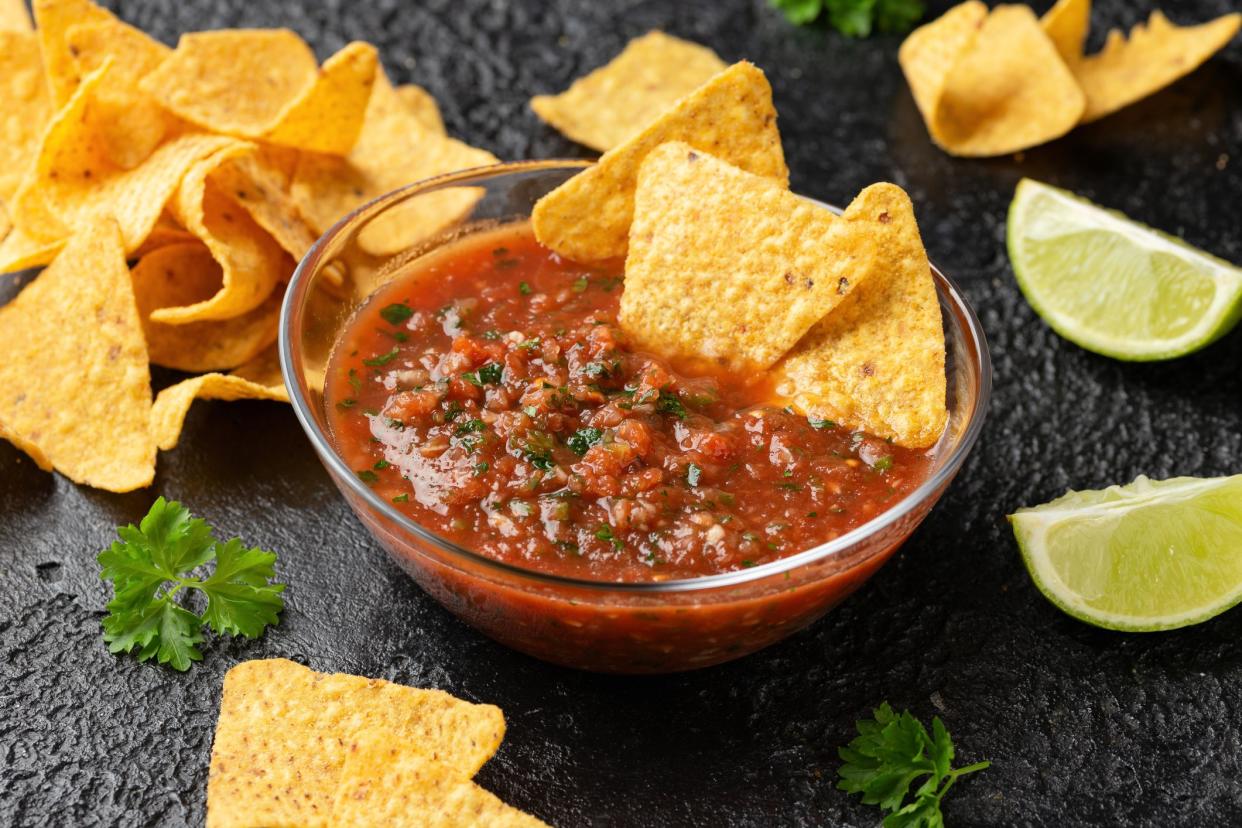 Two homemade chips in a glass bowl of salsa, surrounded by more homemade chips and two slices of lime on a slab of slate