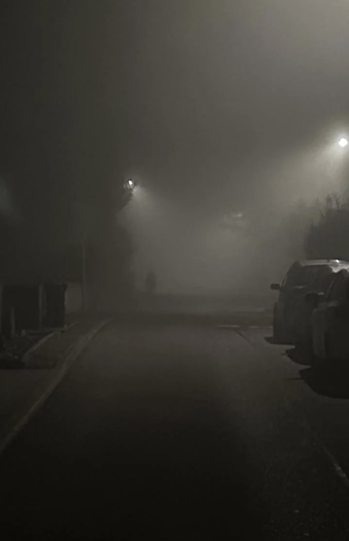 A foggy street at night with streetlights visible and cars parked on the side
