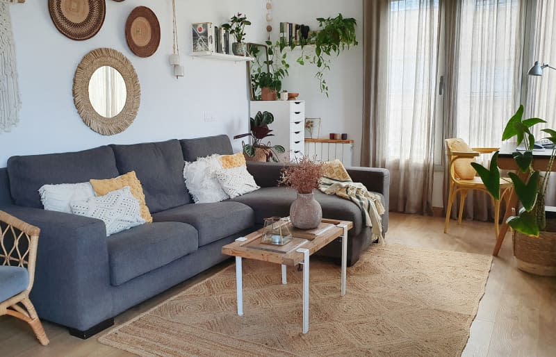 living room with gray sofa, natural fiber rug, wood coffee table with white metal legs, baskets, mirror with straw frame on wall behind sofa, cane armchair with gray cushion, gray armchair on other side of sofa, plants on floating shelf, lots of white, wood, natural accents