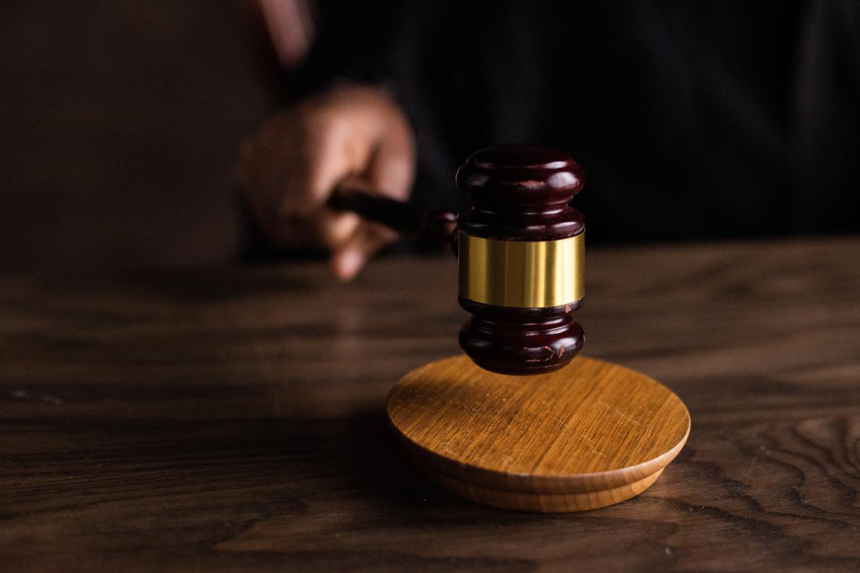 A brown wooden gavel is struck against a hardwood sound block atop a table by a hand jutting out of a black robe.