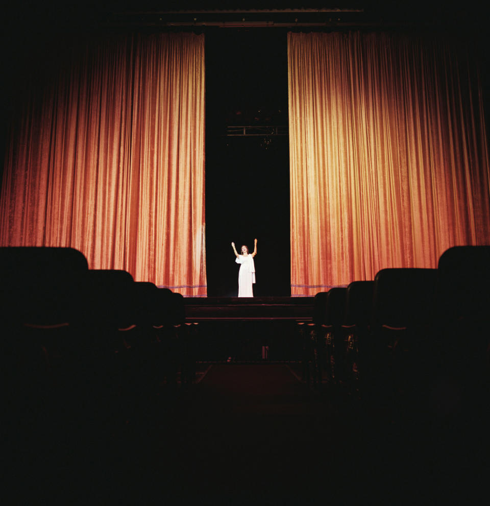 Person on stage with arms raised, audience seats in foreground, theater setting