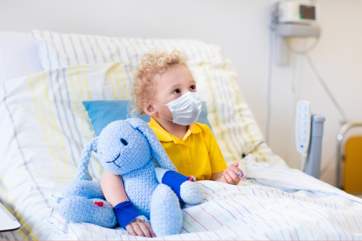 Little boy playing wearing face mask with his toy in bed in hospital room. Child with IV tube and pulse oximeter in clinic. Kid recovering from coronavirus. Corona virus outbreak. Patient in kids ward