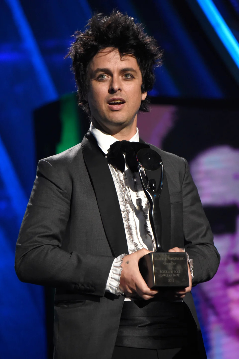 Billie Joe Armstrong holding an award, dressed in a suit with a ruffled shirt, at a formal event