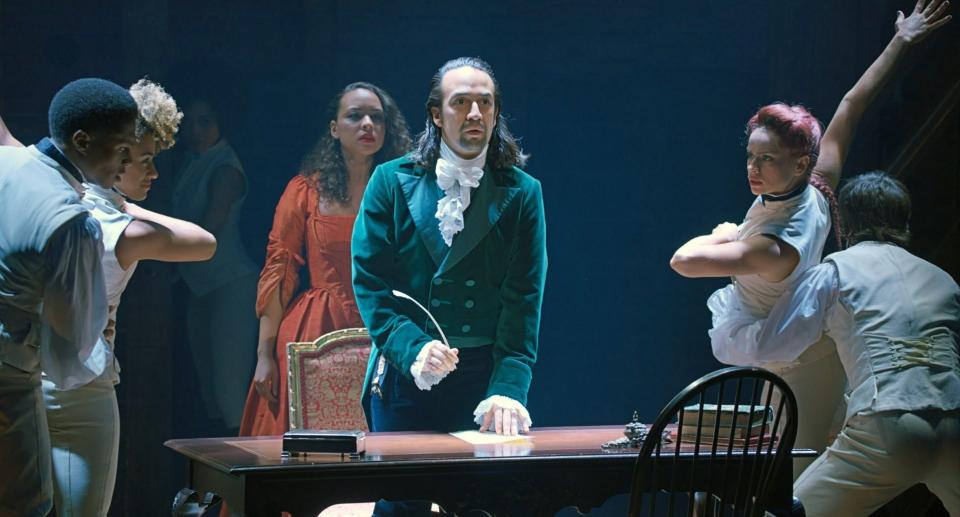Lin-Manuel Miranda stands in front of a desk with a quill while dancers dance around him and Jasmine Cephas Jones sings behind him