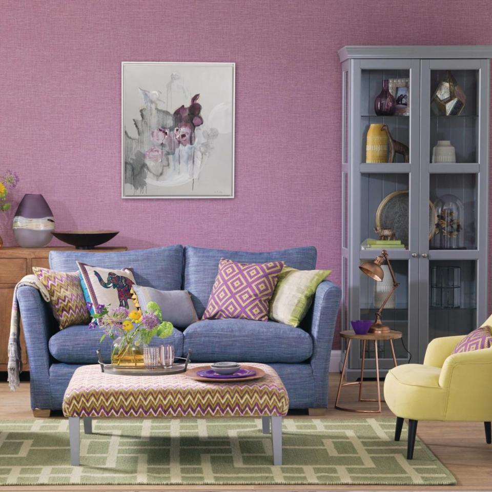 Boho chic living room with mauve wallpaper, cushions on blue Herringbone sofa, turquoise and yellow mid century armchairs and upholstered footstool with zigzag pattern on green rug.