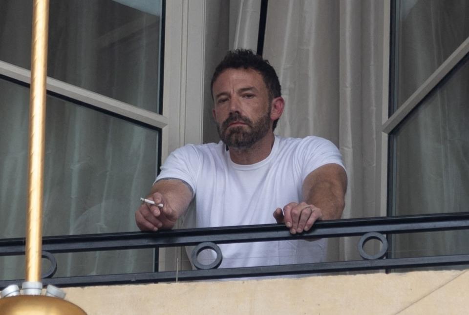 Ben Affleck is on a balcony, leaning on the railing with a cigarette in his hand, looking into the distance.