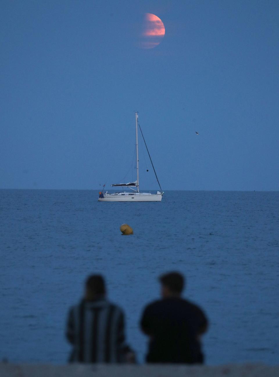 People watch on as the moon rises over a boat moored off of Avon beach in Dorset (PA)