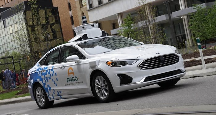 A white Ford Fusion sedan with visible self-driving sensor hardware and Argo AI logos on the doors, driving on a city street.