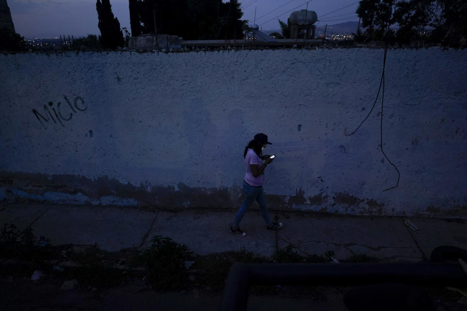 A woman walks along a street at dusk as she looks at her cell phone in Naucalpan, State of Mexico, Mexico, Sunday, July 3, 2022. From January to October in the State of Mexico, there were 120 femicides, which are cases of women killed because of their gender. (AP Photo/Eduardo Verdugo)