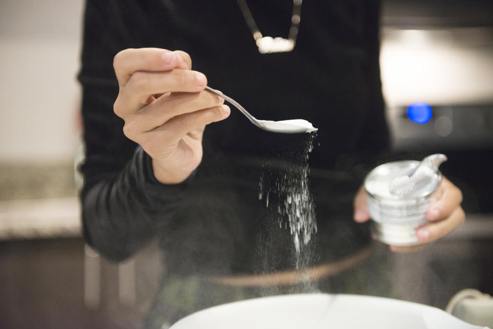 A photograph of a young 20 year old Millennial woman baking cupcakes from scratch at home in the kitchen in Orlando, Florida, USA. She measures out a spoon full of baking soda to add to the bowl of batter.