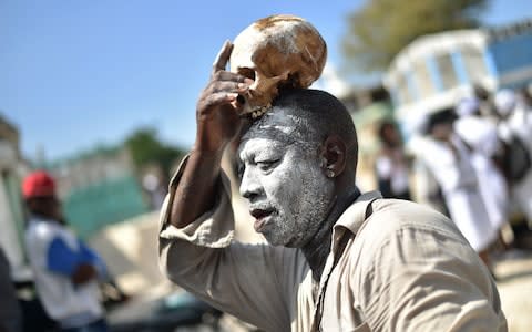 A voodoo devotee with a skull on top of his head is seen during ceremonies honoring the Haitian voodoo spirit of Baron Samdi and Gede on the Day of the Dead in the Cementery of Cite Soleil, in Port-au-Prince, Haiti on November 1, 2017 - Credit: AFP