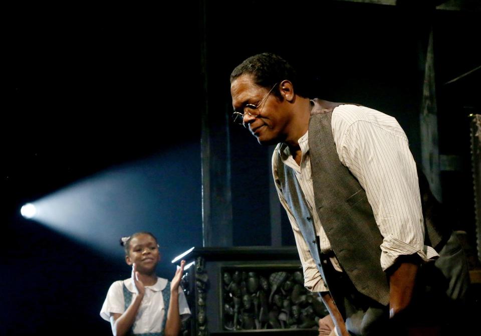 samuel l jackson, wearing a white shirt, olive vest, and glasses, bows on a stage, while a young girl claps in the background