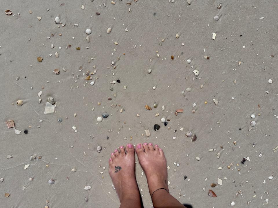 Feet with pink painted toenails on a beach with several shells and shell pieces in white sand