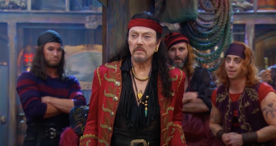 actor Christopher Walken wears a red coat as Captain Hook and and stands in front of three men dressed as pirates